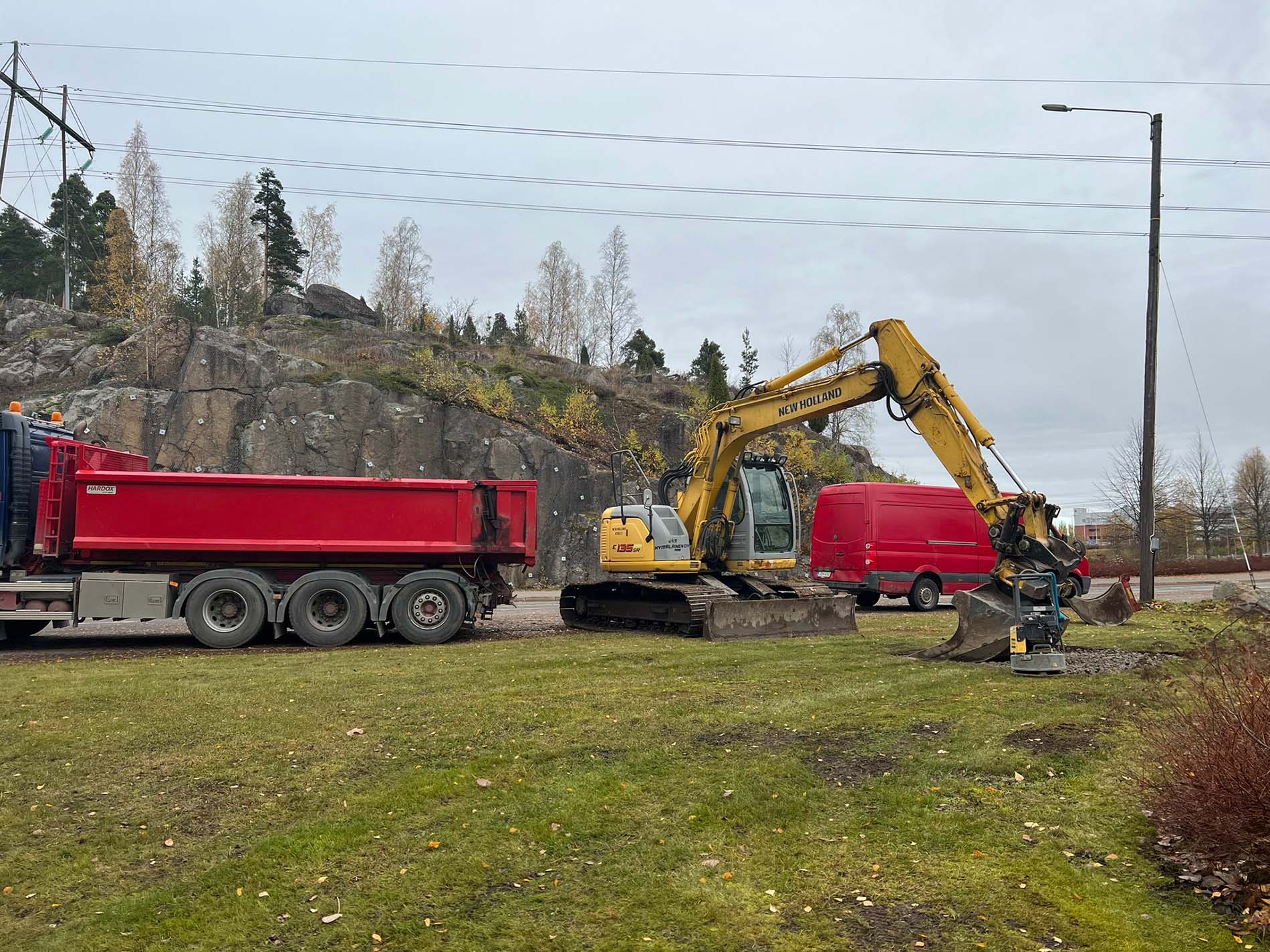 Keltainen New Holland -kaivuukone ja punainen pakettiauto työmaalla